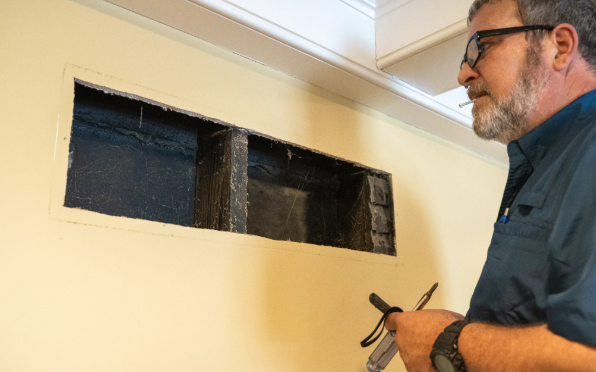 Man inspecting a dark wall-mounted panel beneath a ceiling vent inside a room