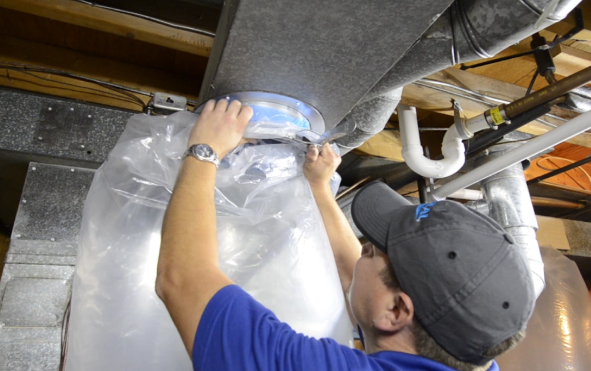 Worker in blue shirt inspecting and sealing ductwork under a ceiling with foil insulation.