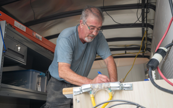 Technician working inside an equipment compartment, leaning over wires and tools in a narrow metal space