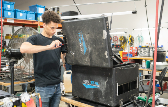 Person inspecting a large black machine in a workshop cluttered with tools and equipment