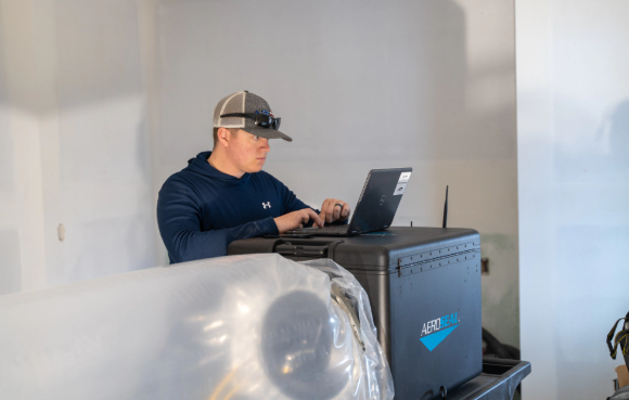 Person in a cap uses a laptop on a black equipment case in a room with white walls and wrapped furniture.