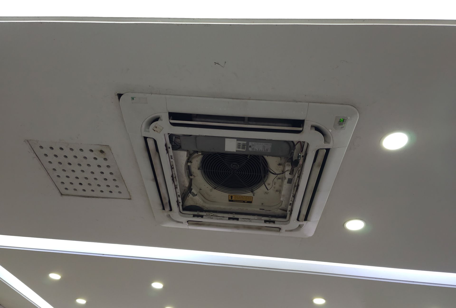Ceiling-mounted square air vent or HVAC unit with surrounding recessed lights in a white interior ceiling