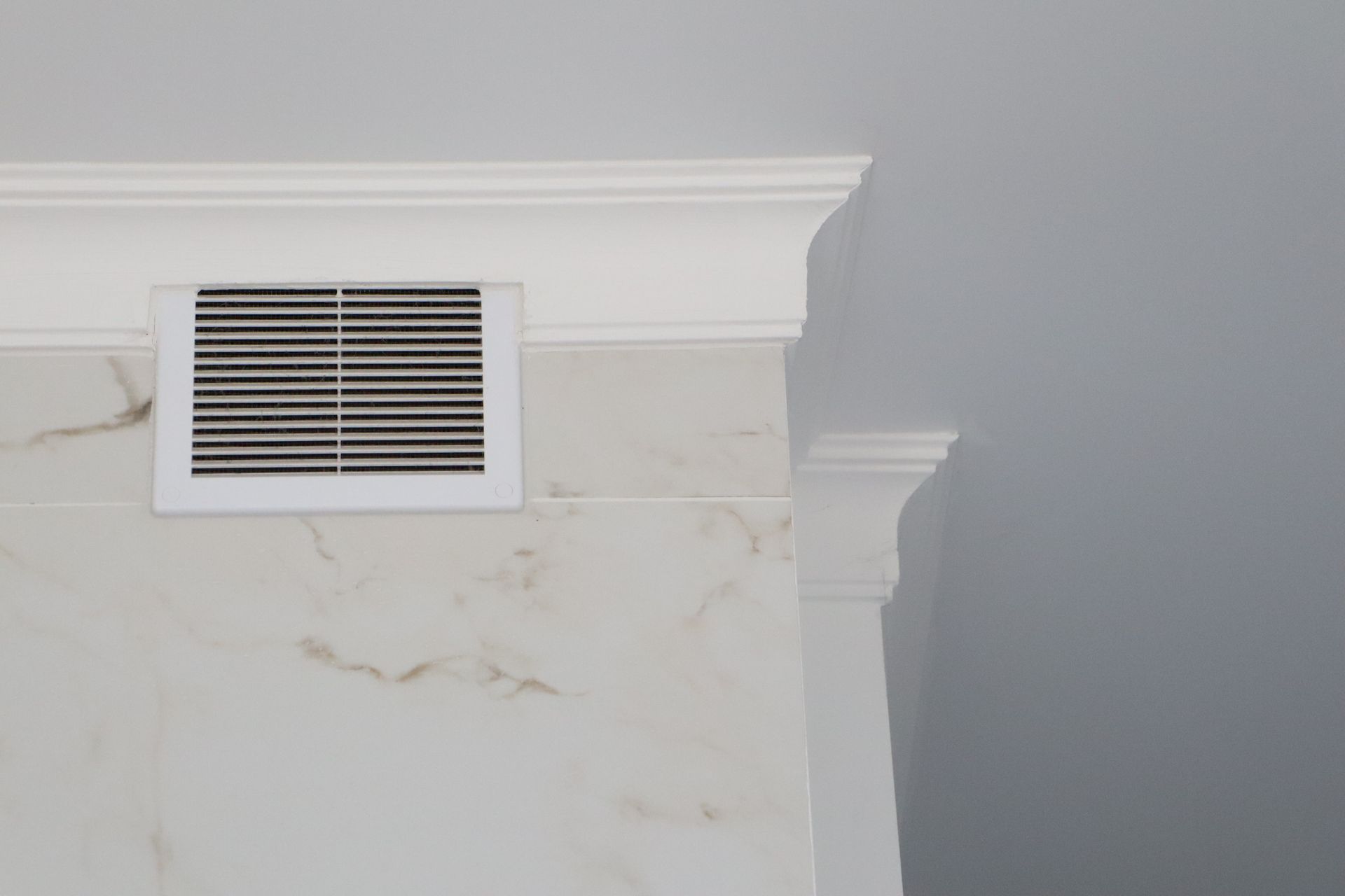 White wall vent beneath crown molding on a light gray wall with marble-patterned trim
