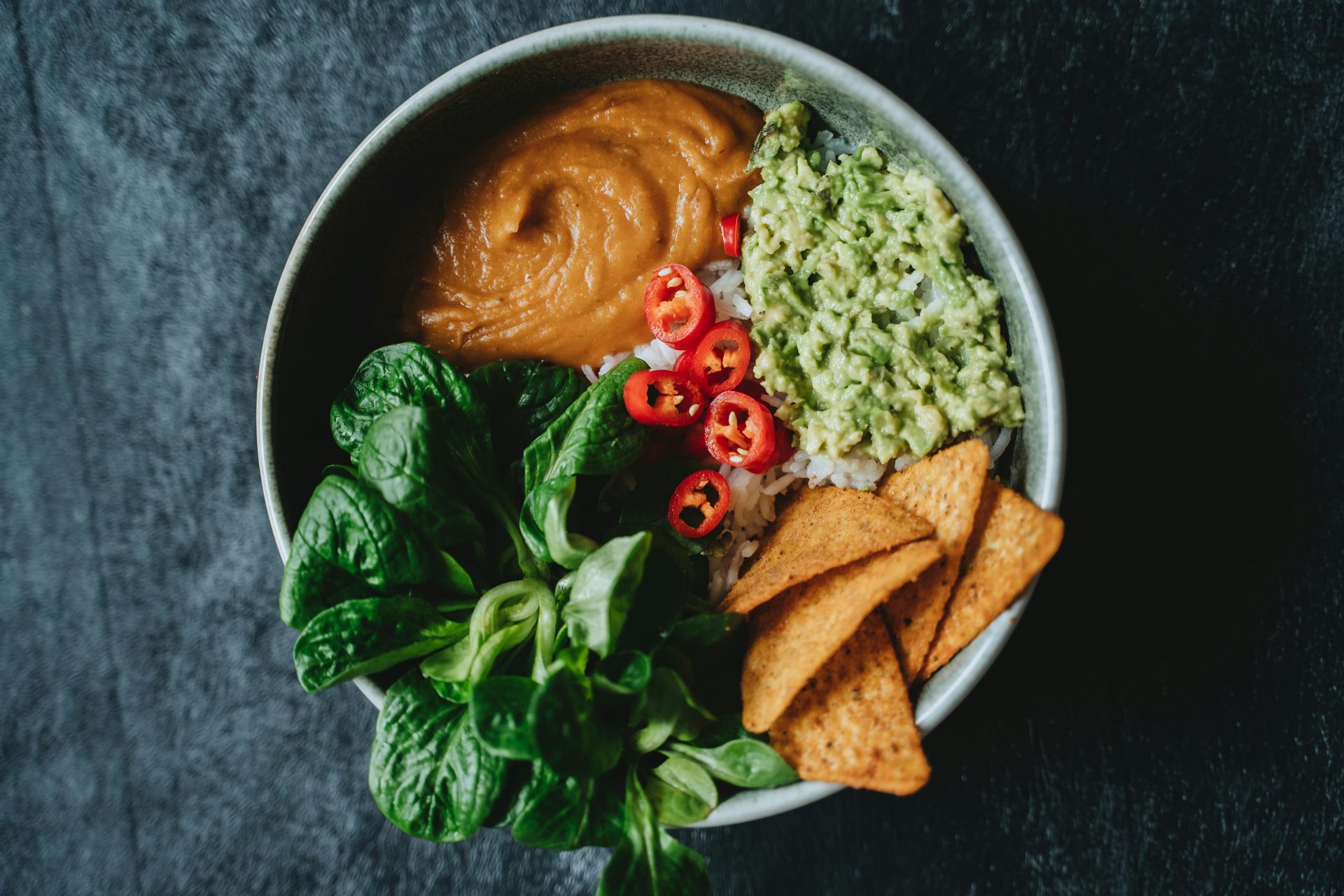 A bowl of food with guacamole , rice , tortilla chips and spinach on a table.