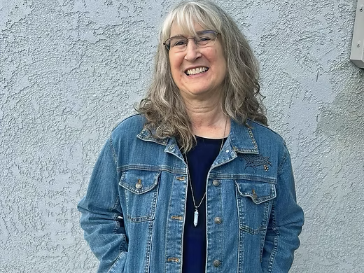 A smiling person with long gray hair wears a blue denim jacket over a dark top and a crystal pendant against a gray wall.