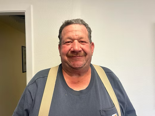 A person with a mustache smiling in a blue shirt with beige suspenders against a plain white wall.