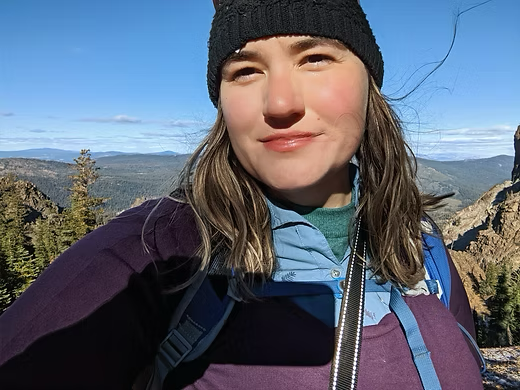 Person wearing a beanie and purple top poses for a selfie on a sunny mountain ridge with pine trees and blue sky.