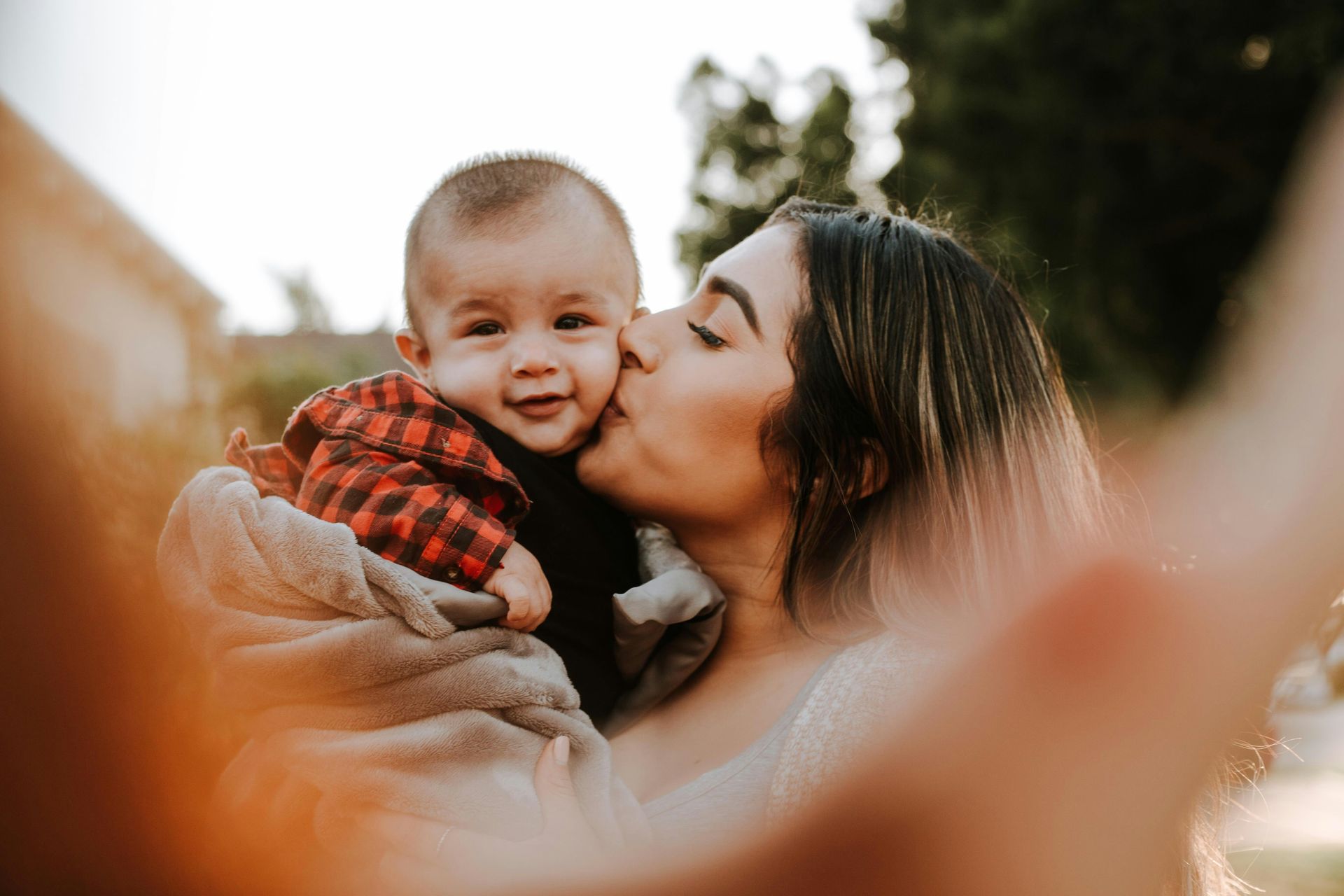 A person kisses a baby on the cheek while holding them in a soft, gray blanket outdoors.