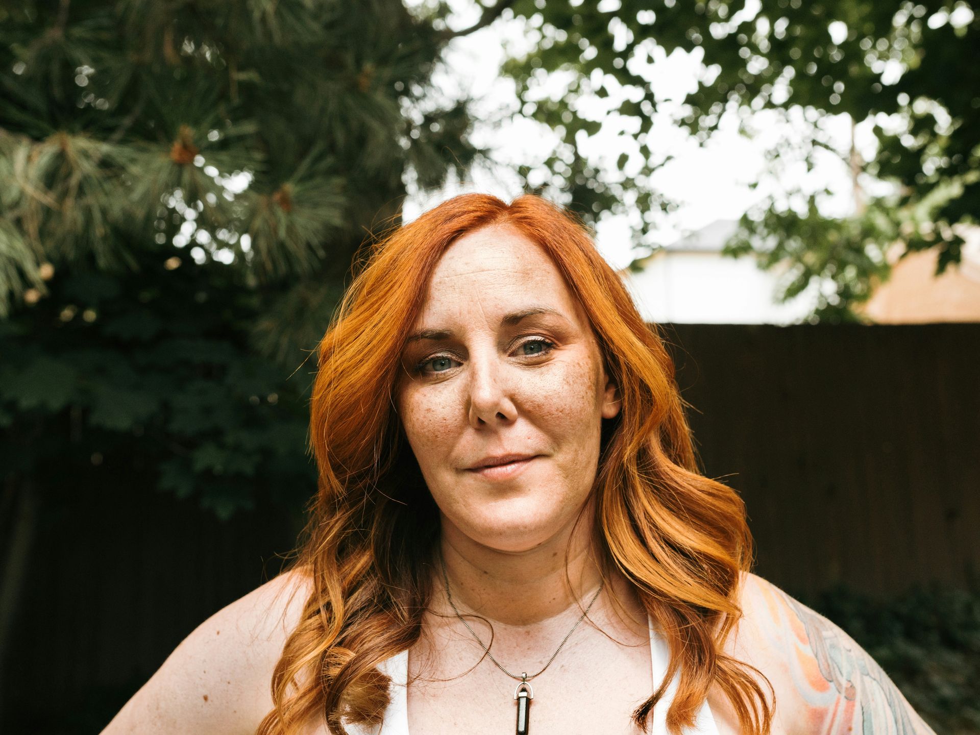 A person with shoulder-length wavy red hair wears a necklace while standing outdoors in front of a tree and wooden fence.