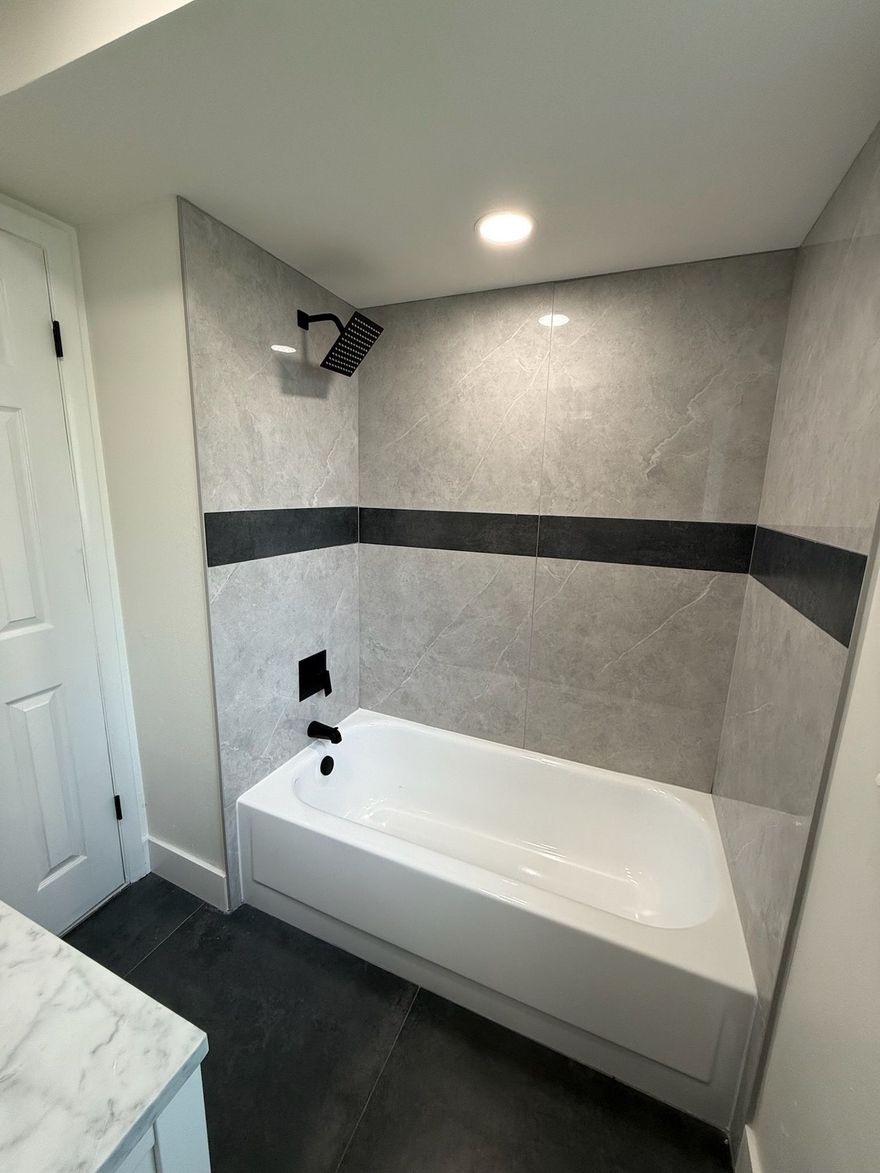 A newly renovated bathroom with a white tub, gray and black tiled shower, and black fixtures.