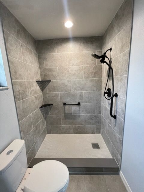 Accessible shower with gray tile, grab bar, and showerhead, next to a toilet.