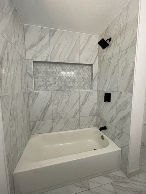 Bathroom with marble-patterned tiles, bathtub, shower head, and recessed shelf with gray mosaic tiles.