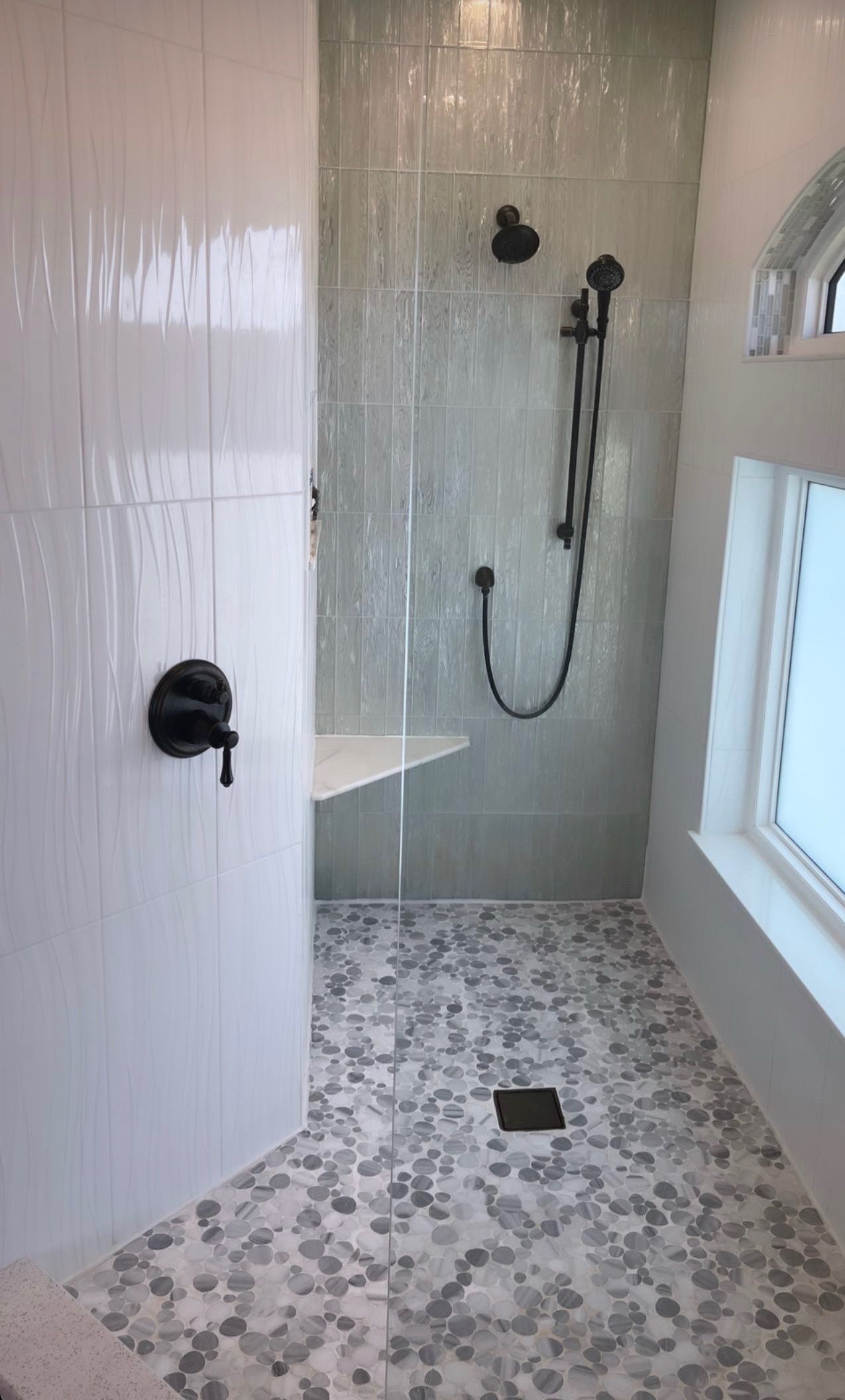 Modern tiled shower with glass door, dark fixtures, and a corner bench.