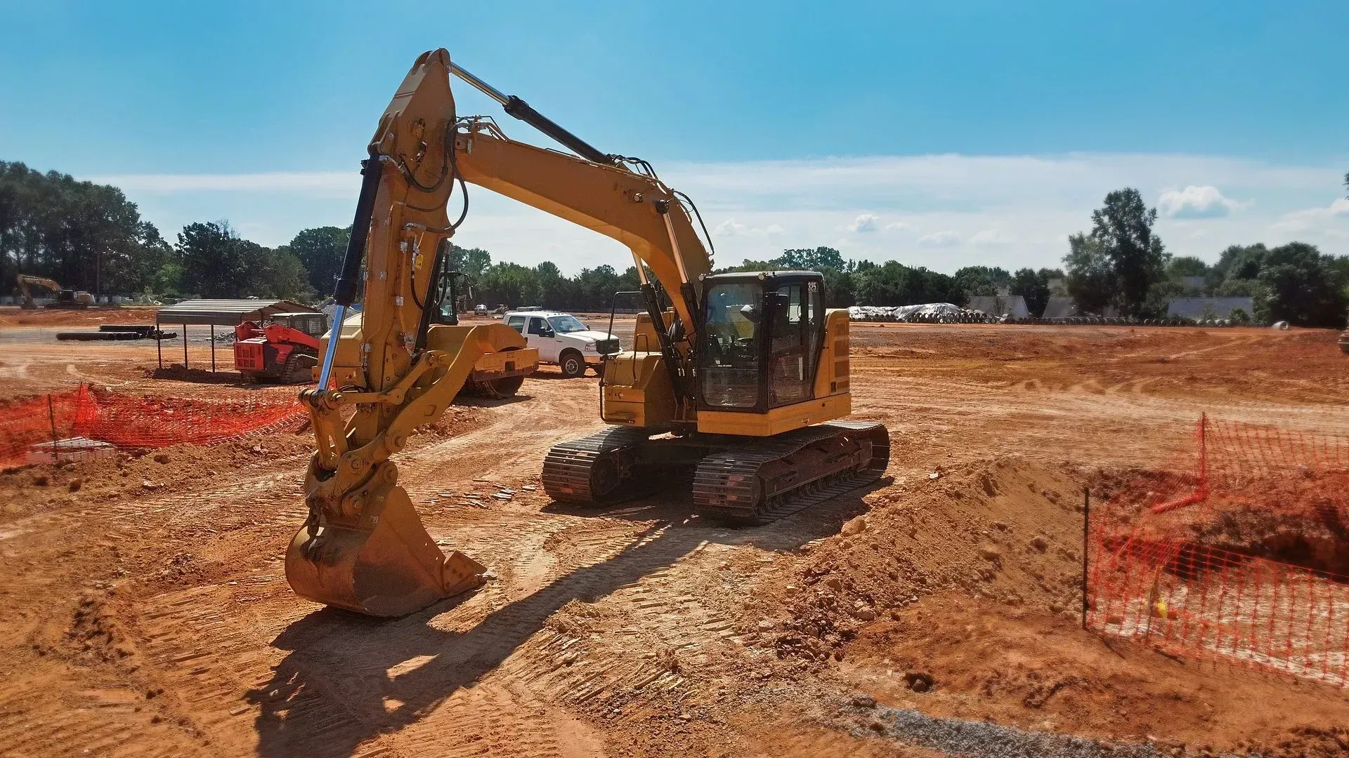 Yellow excavator working on a dirt construction site with orange safety fencing and trees in the background