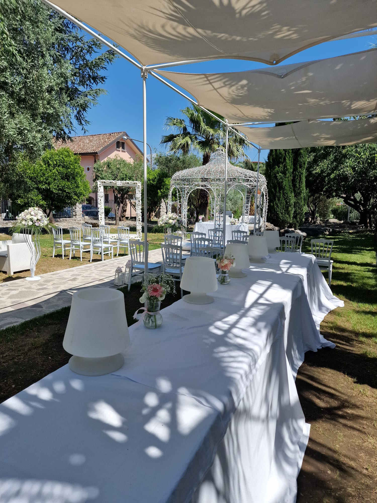 Un lungo tavolo con tovaglie bianche e lampade sotto un baldacchino in un parco.