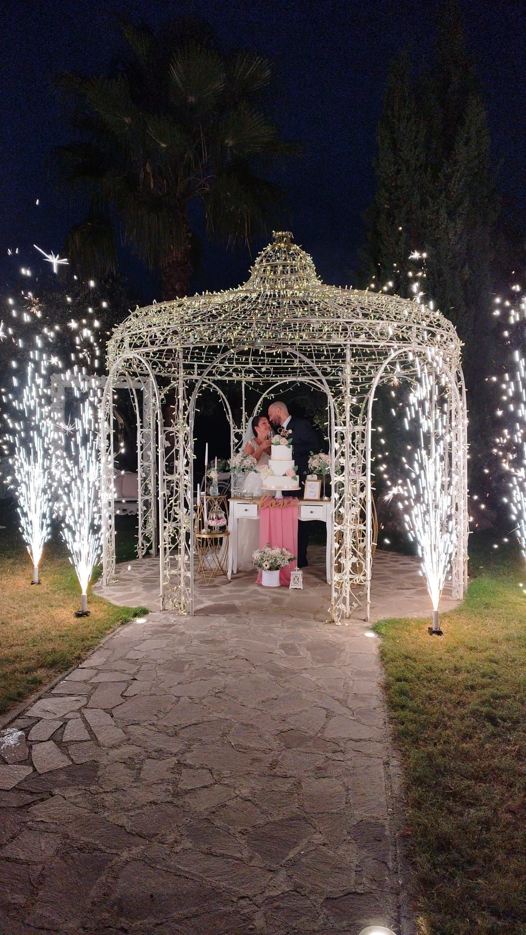 Una sposa e uno sposo sono in piedi sotto un gazebo con fuochi d'artificio sullo sfondo.