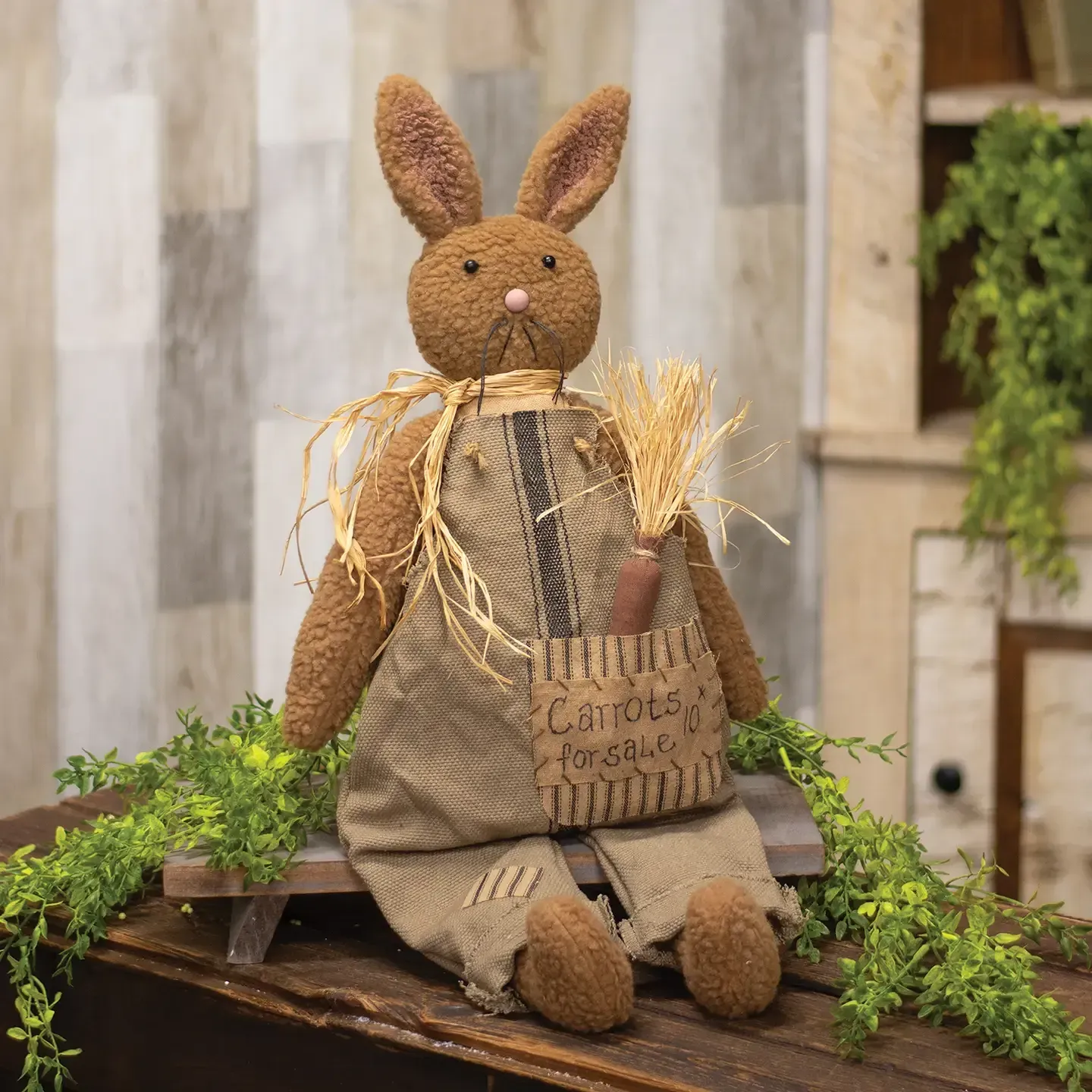 A primitive-style fabric bunny doll in burlap overalls and a straw collar, holding a carrot, sitting on a wooden stool.