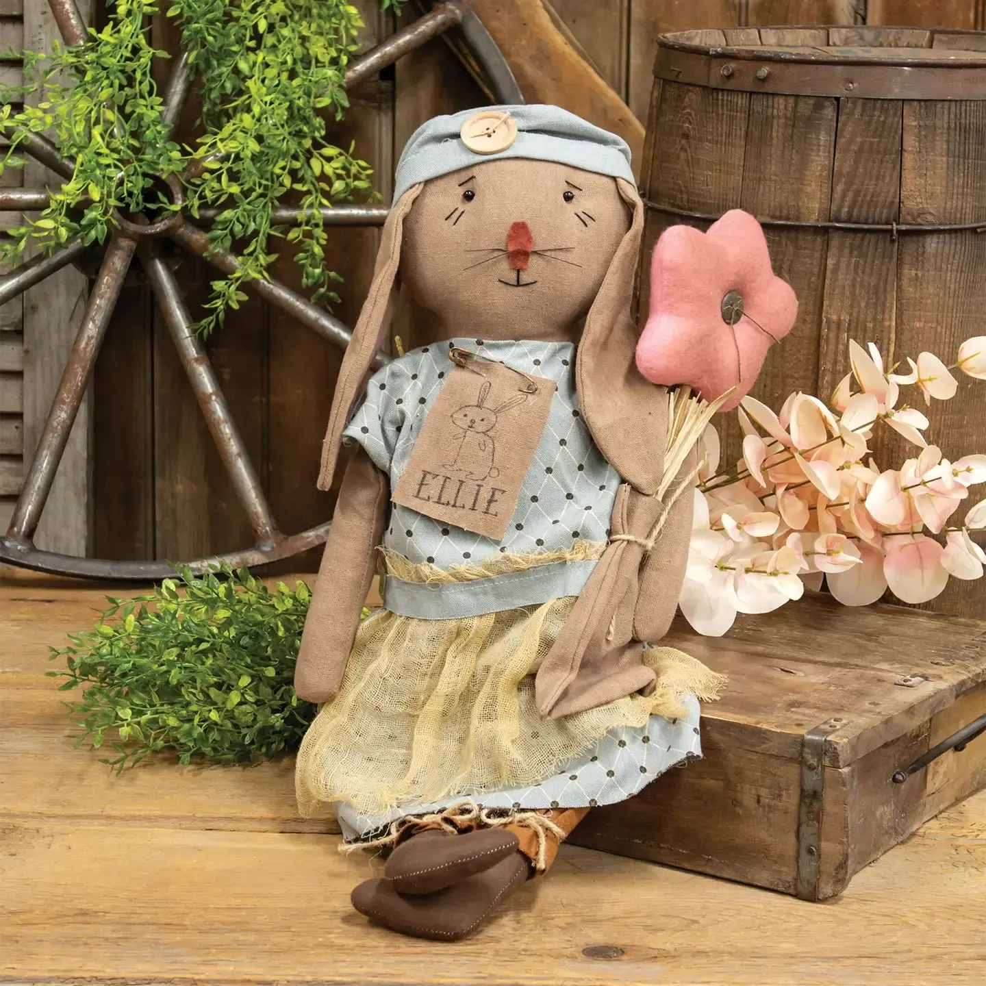 A rustic fabric bunny doll in a polka-dot dress and blue headwrap, holding a pink fabric flower, sitting by a wood barrel.