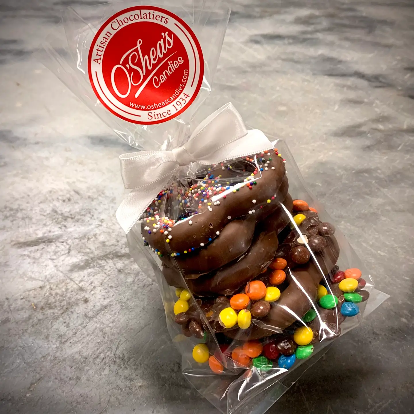 Chocolate-covered pretzel twist with sprinkles and M&Ms, in clear cellophane bag tied with white ribbon; O'Shea's logo.