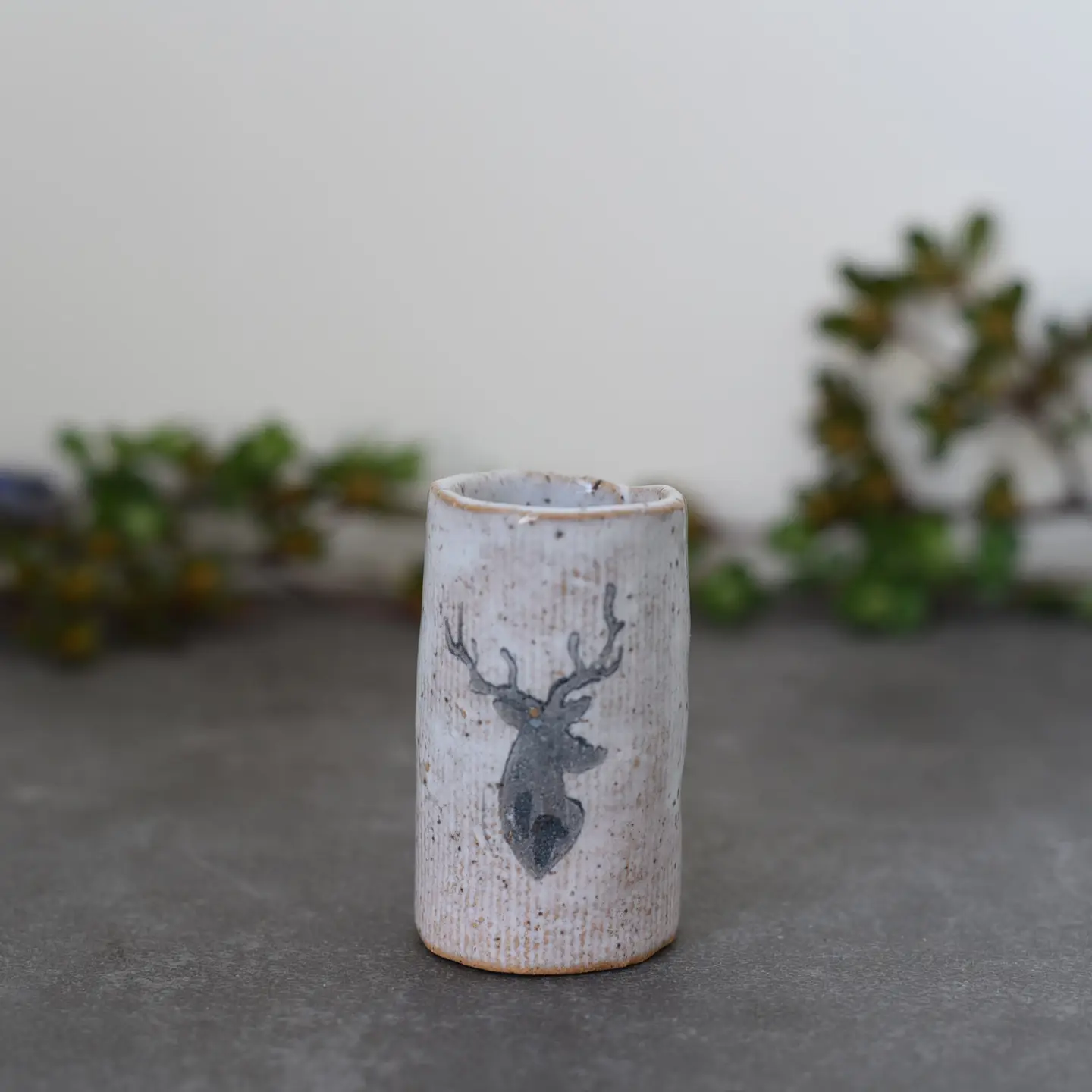 Cylindrical ceramic vase with a deer head illustration. Neutral tones; blurred green foliage background.