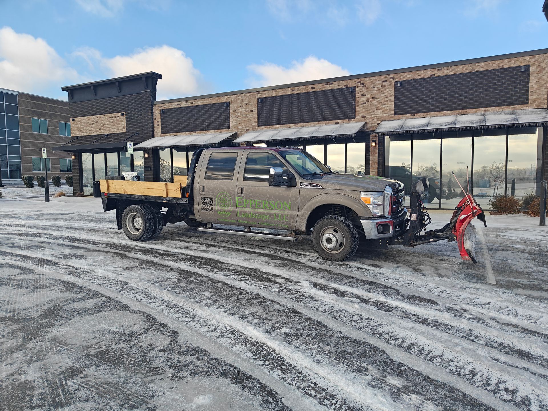A gray truck with a plow on a snowy street in front of a brick building.