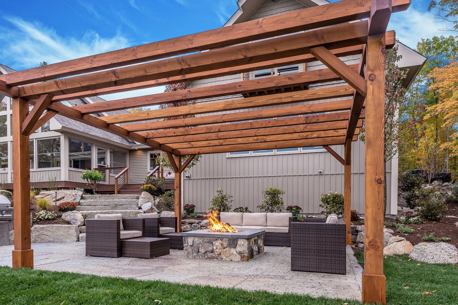 Wooden pergola over a stone patio with seating, a fire pit, and backyard view.