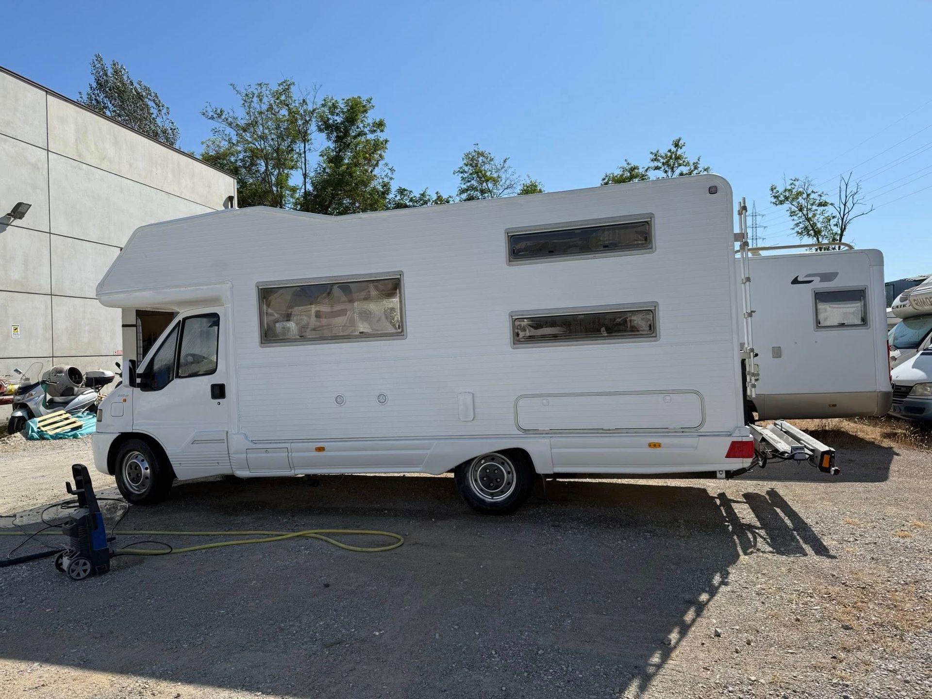 Un camper bianco parcheggiato all'aperto sotto un cielo azzurro. Sono visibili un altro camper e un edificio.
