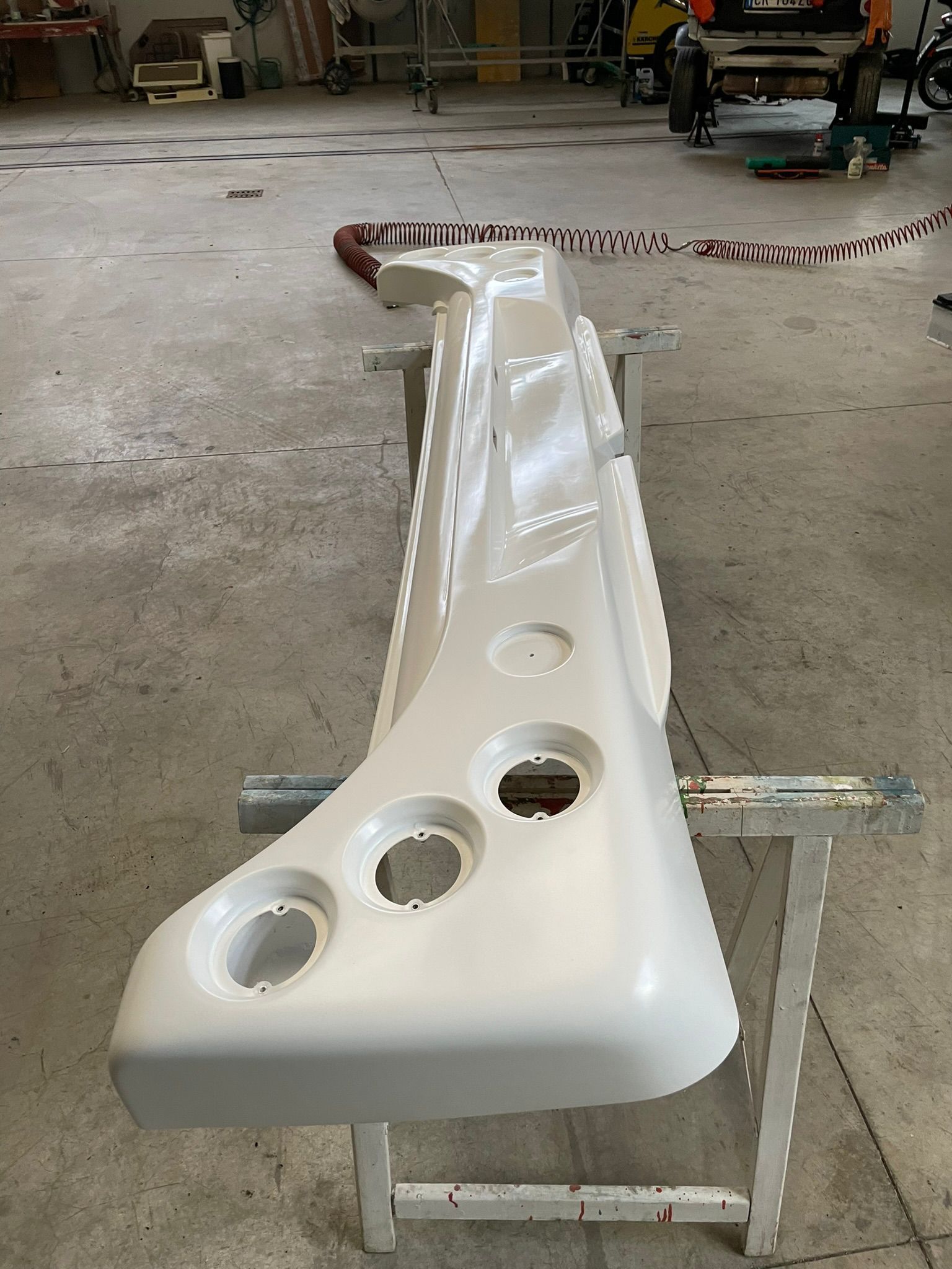 Paraurti bianco con tre grandi fori, appoggiato su cavalletti in un'officina.
