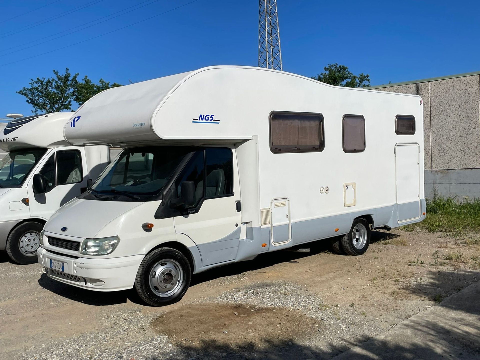 Camper bianco e grigio parcheggiato sulla ghiaia, con un cielo azzurro sullo sfondo.