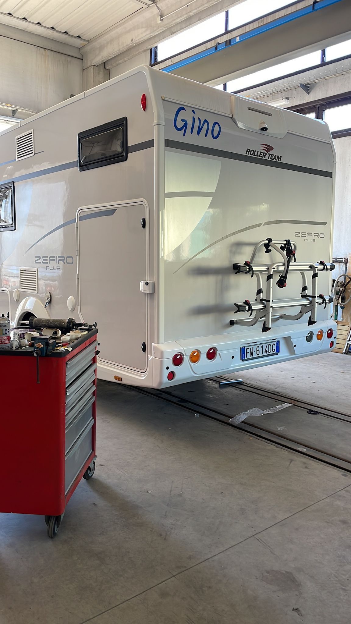 Camper bianco in un garage con una cassetta degli attrezzi rossa in primo piano e un portabiciclette sul retro.