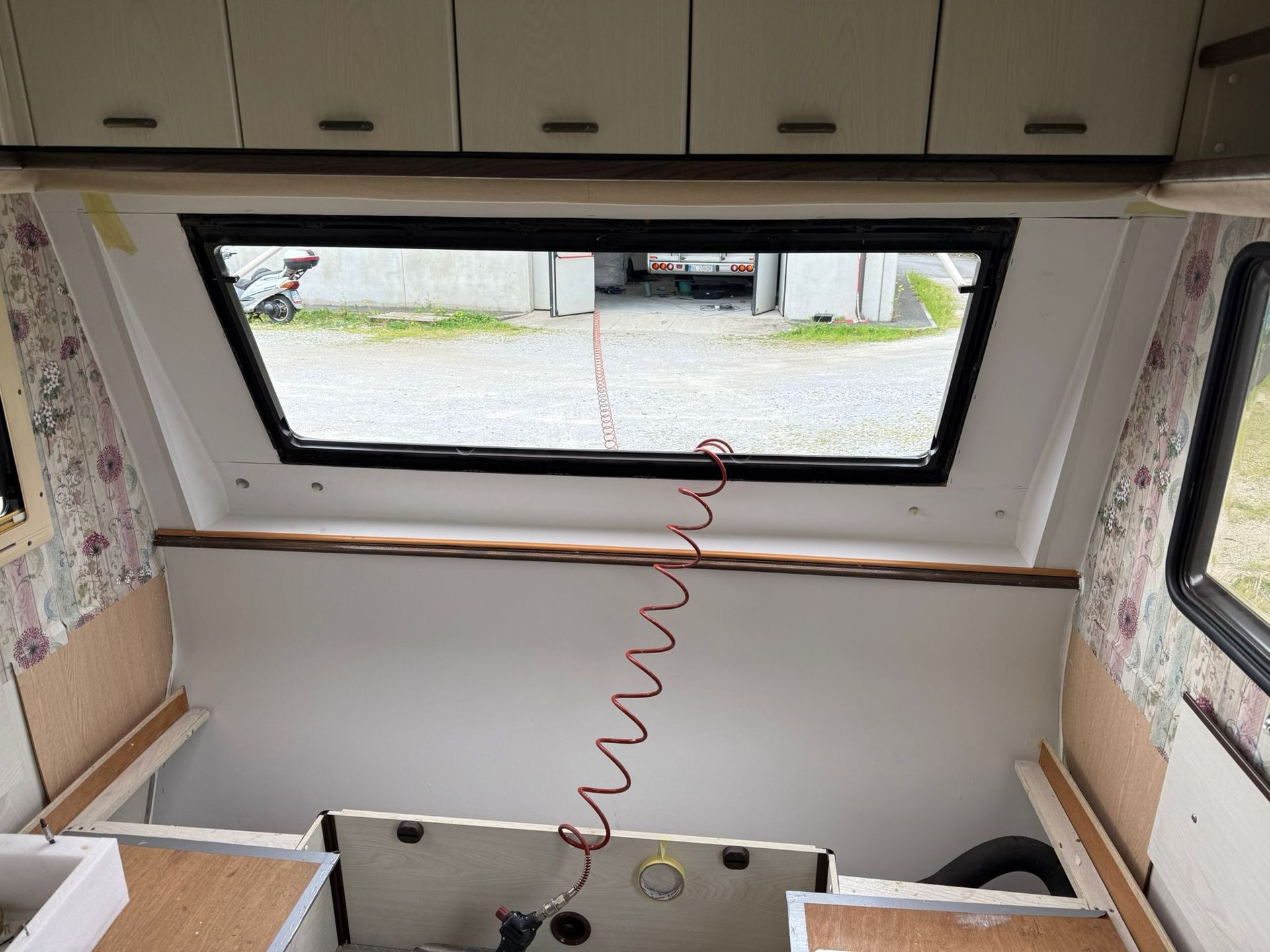 Interno del camper: mobili bianchi, ampia finestra, tubo flessibile rosso a spirale, zona di seduta/stivaggio vuota.