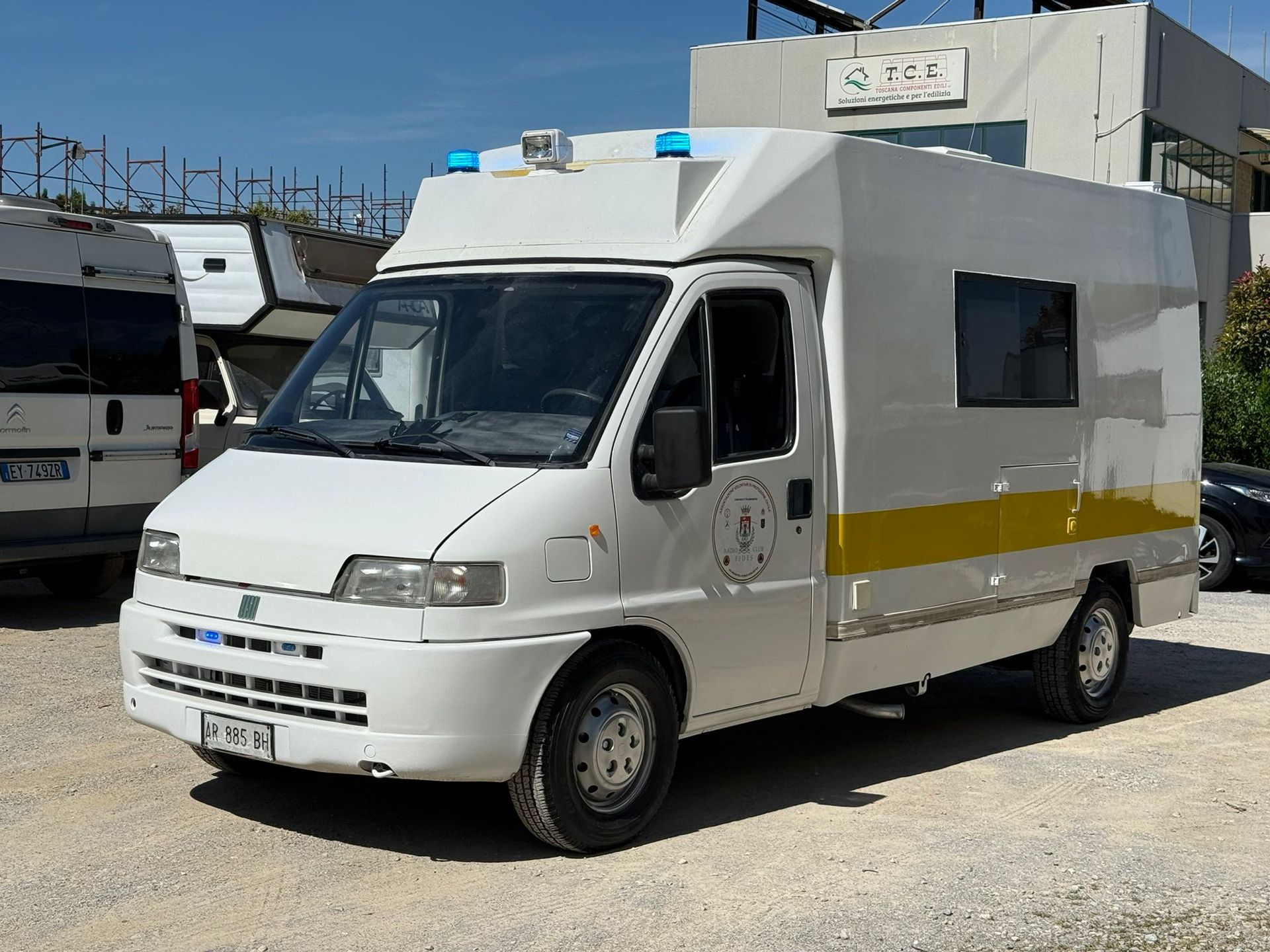 Ambulanza bianca con luci blu, parcheggiata su una superficie ghiaiosa. Striscia gialla lungo la fiancata.