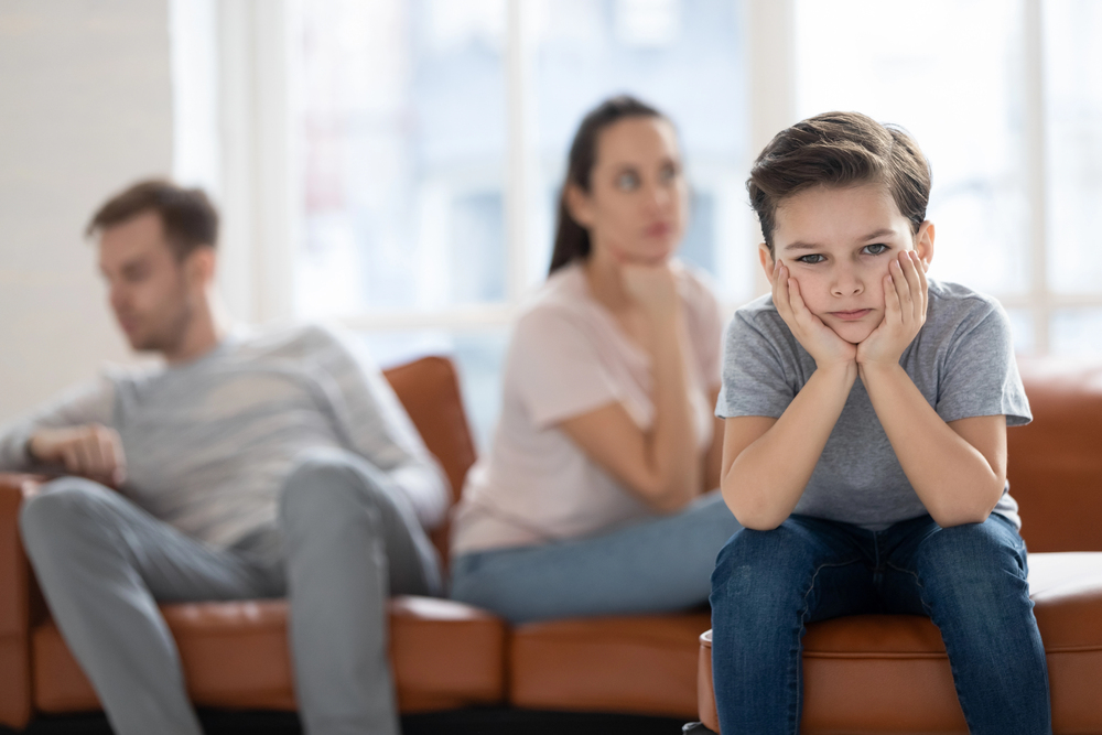 Sad boy with parents in background, sitting on sofa.
