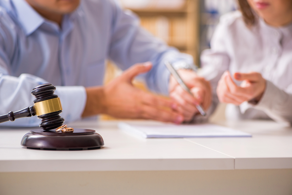 Gavel with wedding ring on table, couple signing documents, possibly a divorce settlement.