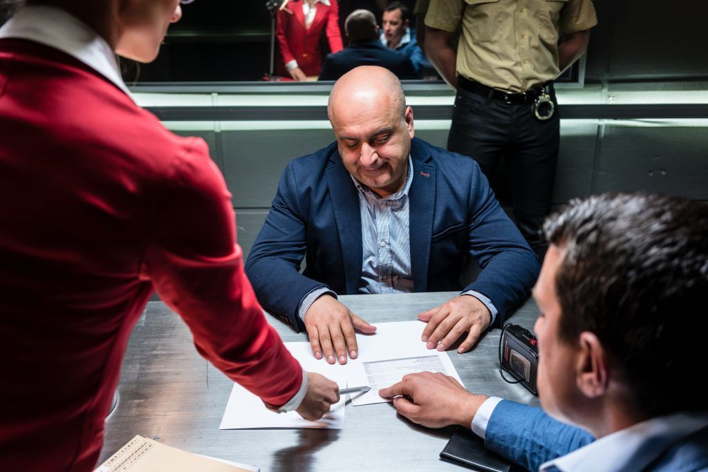 Man in interrogation room being questioned by two people in red suit.