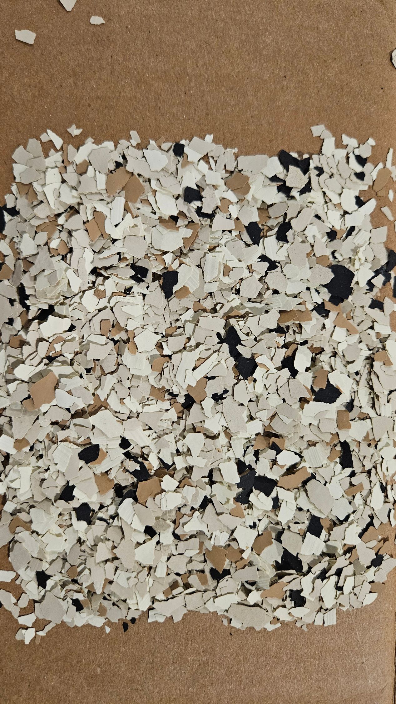 Tan, black, and white decorative epoxy floor flakes on a cardboard surface.
