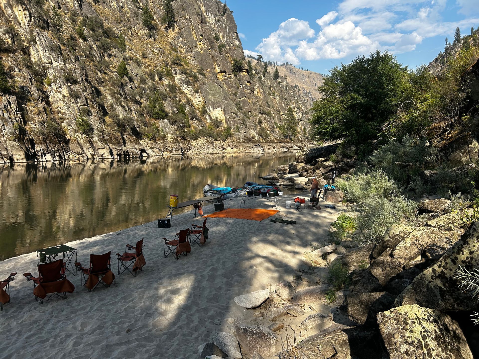A group of people are sitting on a sandy beach next to a river.