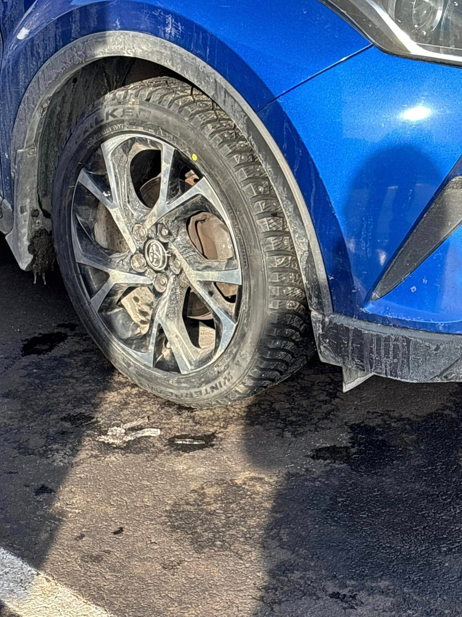 A blue car with a dirty tire is parked in a parking lot.
