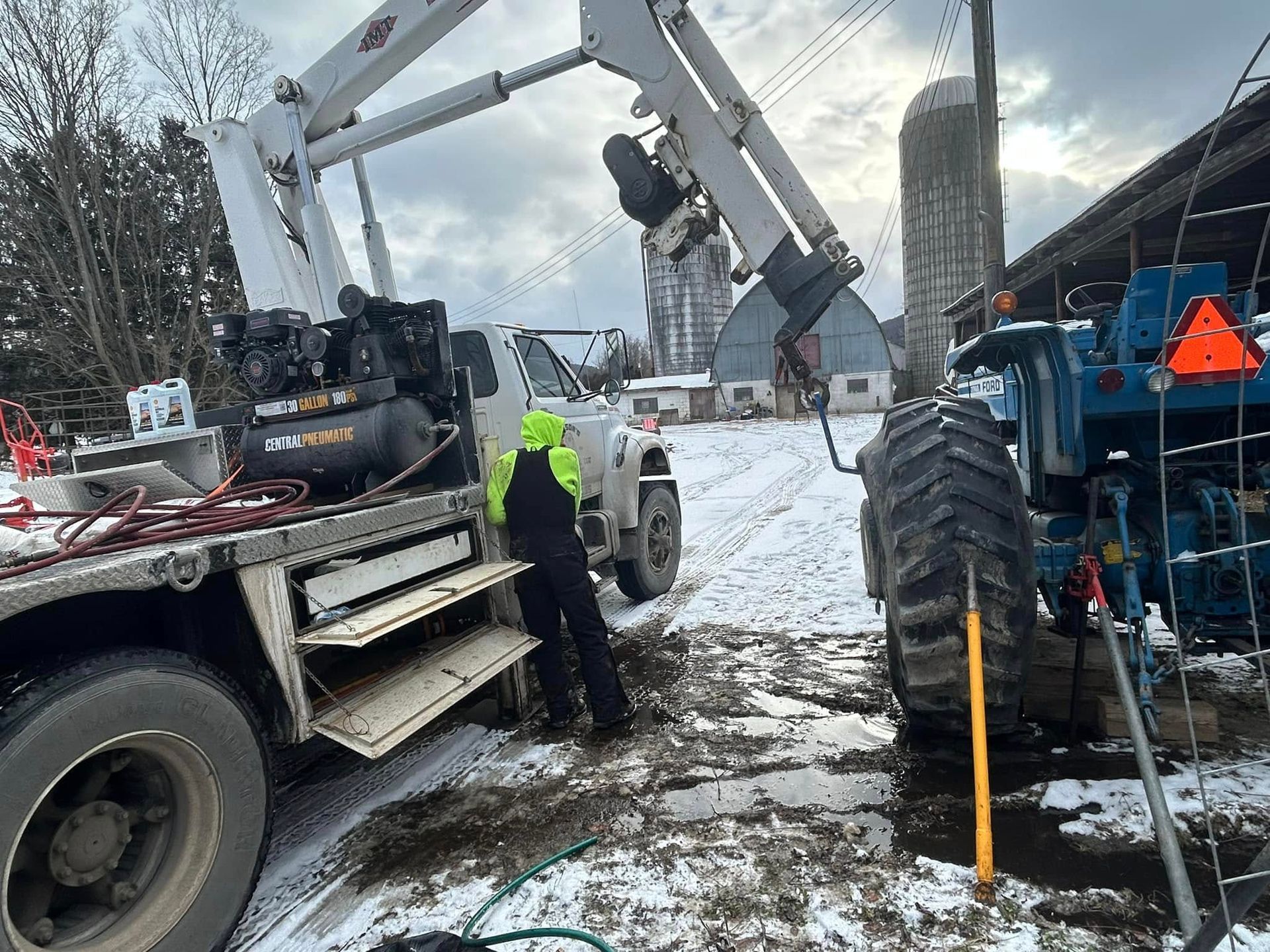 A man is working on a tractor in the snow next to a tow truck.