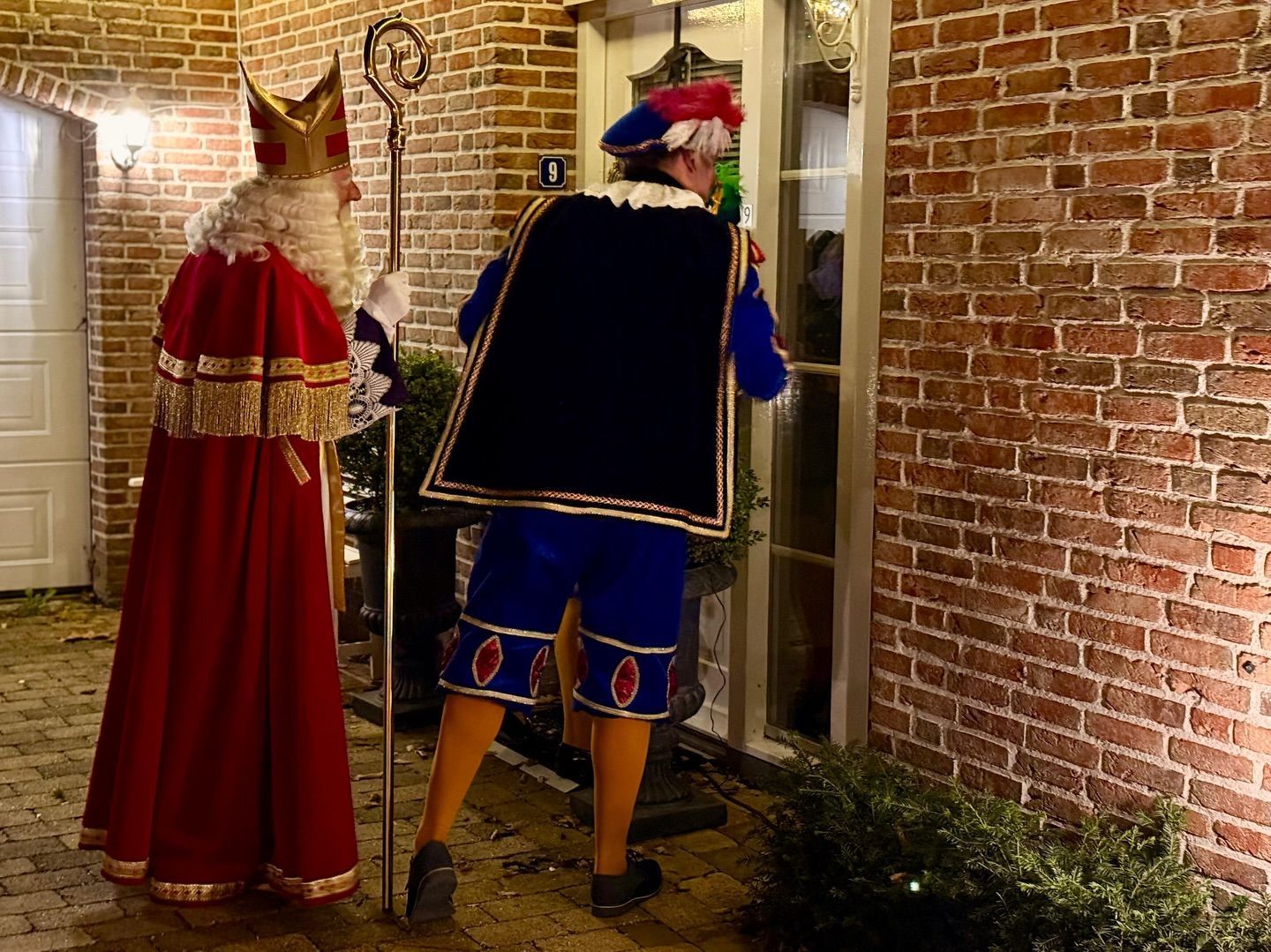 Twee verklede personen staan 's nachts bij een bakstenen muur in een warm verlicht steegje.