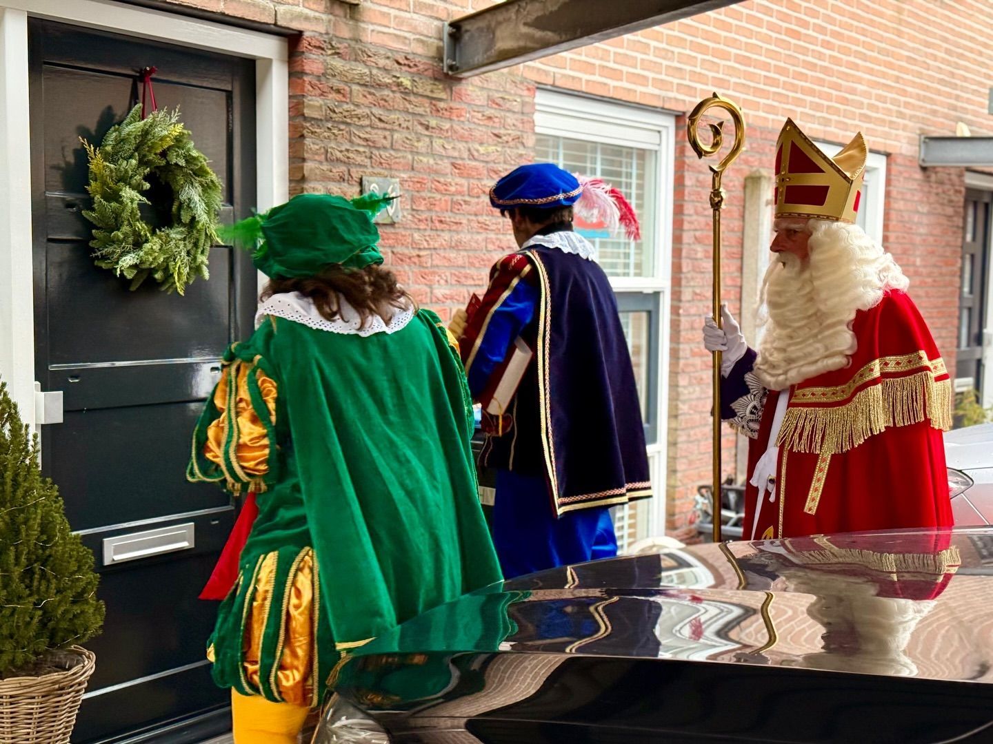 Drie personen in feestelijke mantels en hoeden naderen een huis dat versierd is met een kerstkrans.