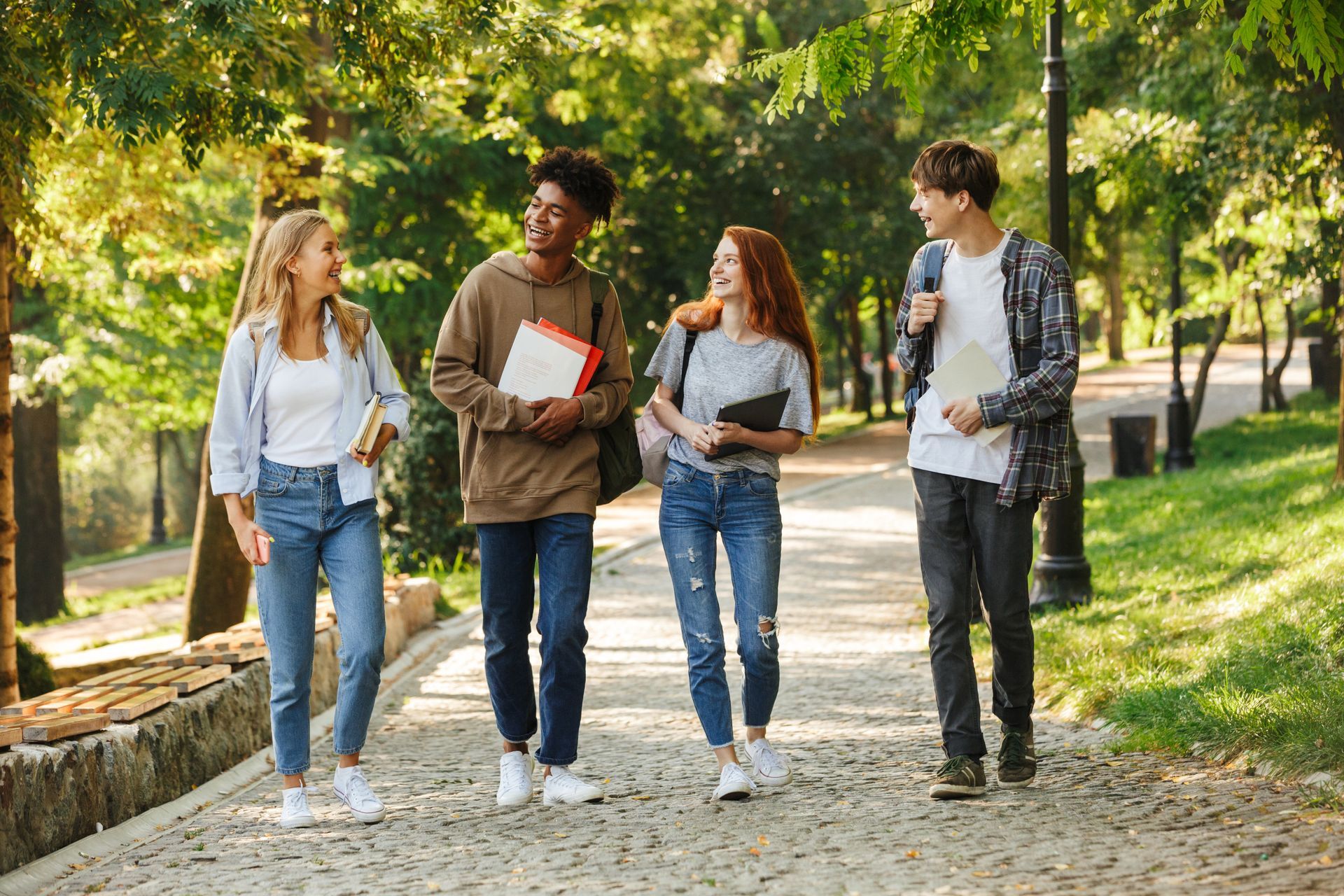 Four students walking and talking on a park path, holding books. Sunlight streams through trees.