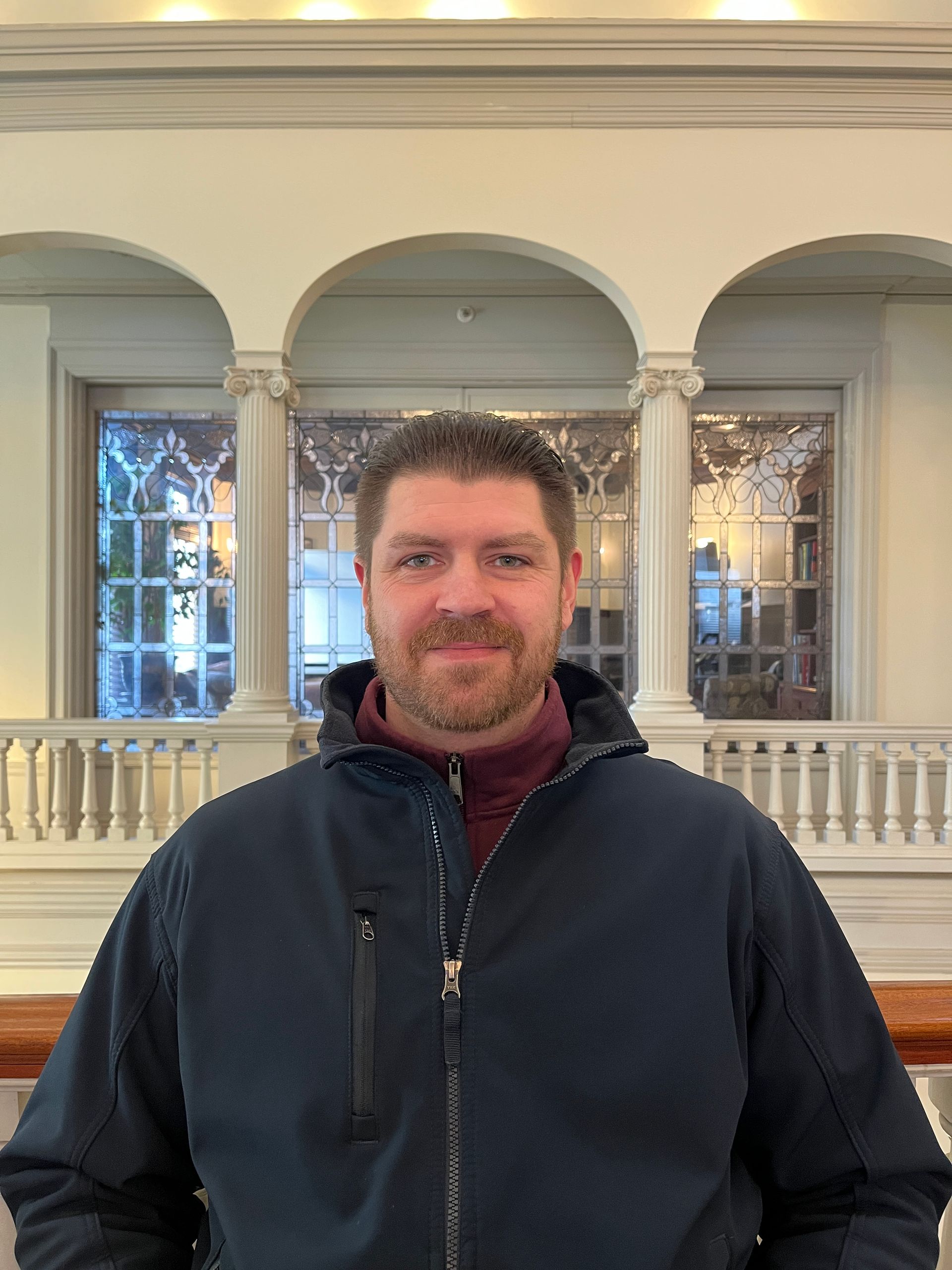 Man in a dark blue jacket smiles, standing inside a building with arches and windows.