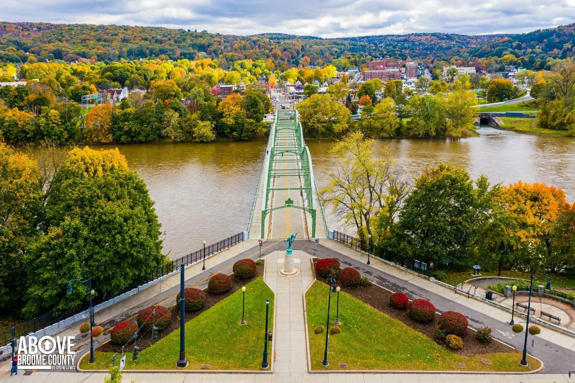 Green suspension bridge over a river, surrounded by fall foliage, town in the background, overcast sky.