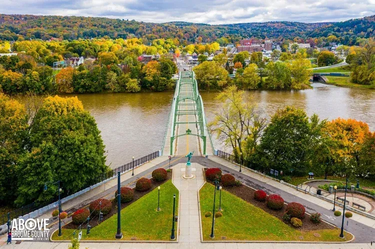 A green suspension bridge over a river, surrounded by fall foliage and a town.
