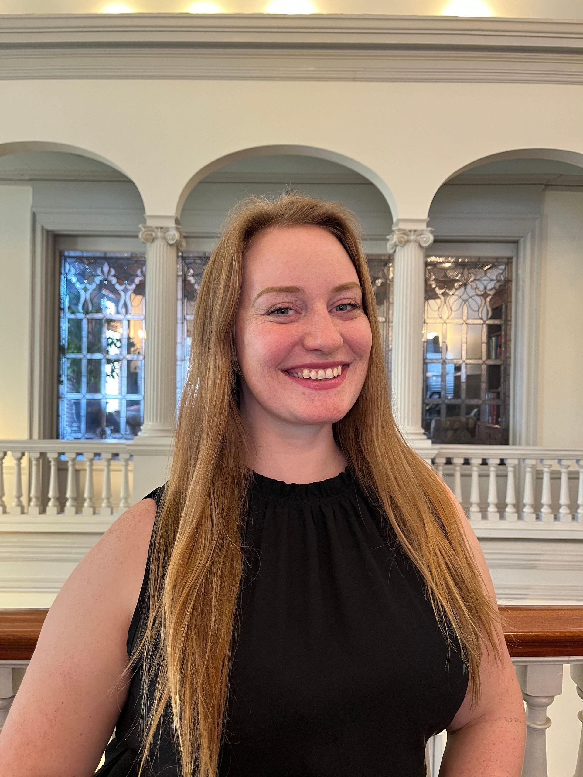 Woman with long blonde hair, smiling, in a black top, in a building with archways and columns.