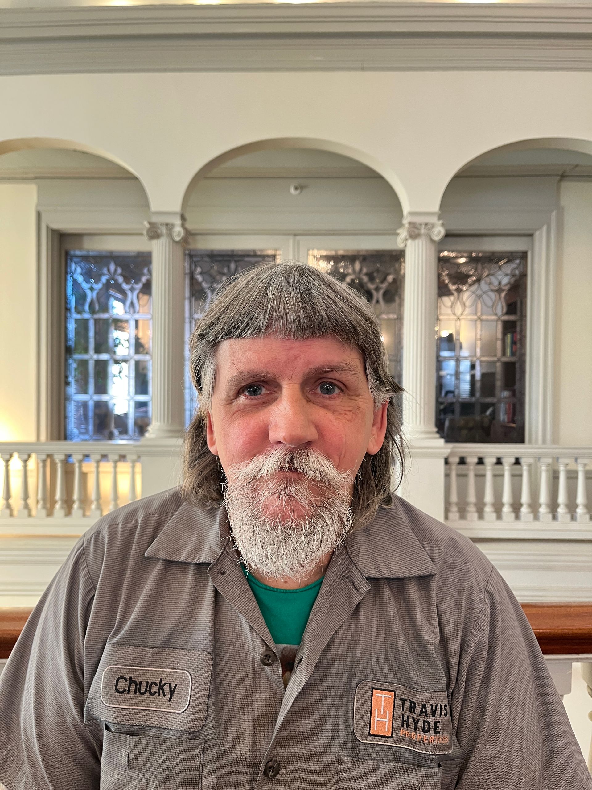 Man with frosted hair and beard in striped shirt stands near a balcony with arched windows.