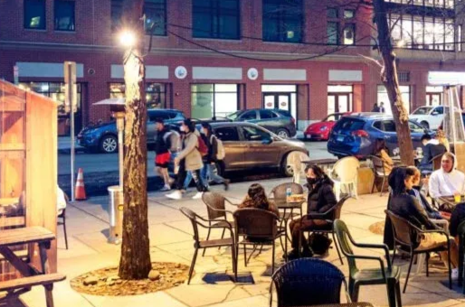 Outdoor cafe at night. People seated at tables. Cars parked on the street. Buildings in the background.