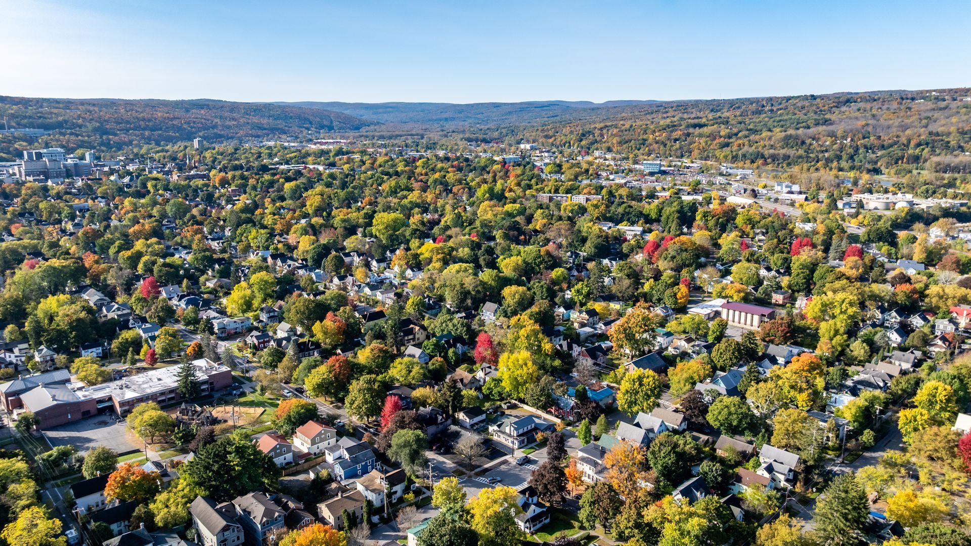 Aerial view of a town in autumn, houses nestled among colorful trees under a clear blue sky.