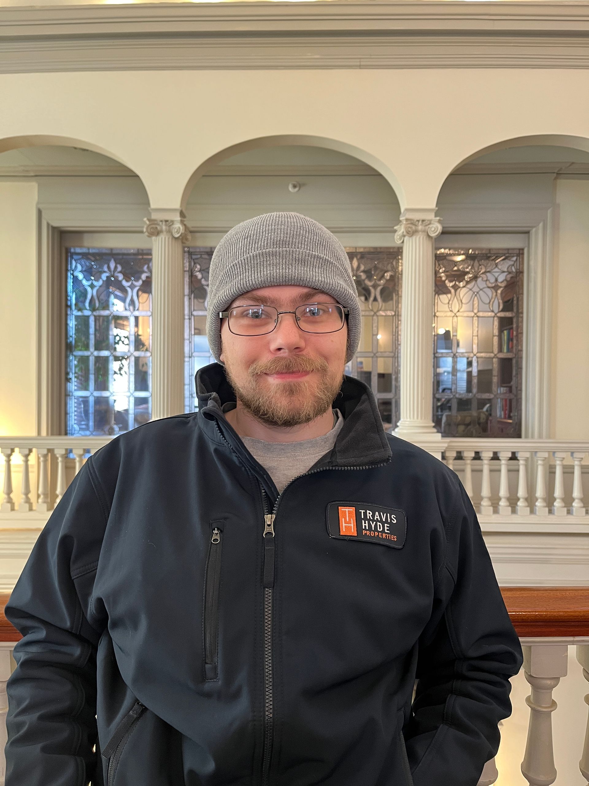 Man wearing a gray beanie and glasses, navy jacket. Standing in a building with arched windows and a white railing.